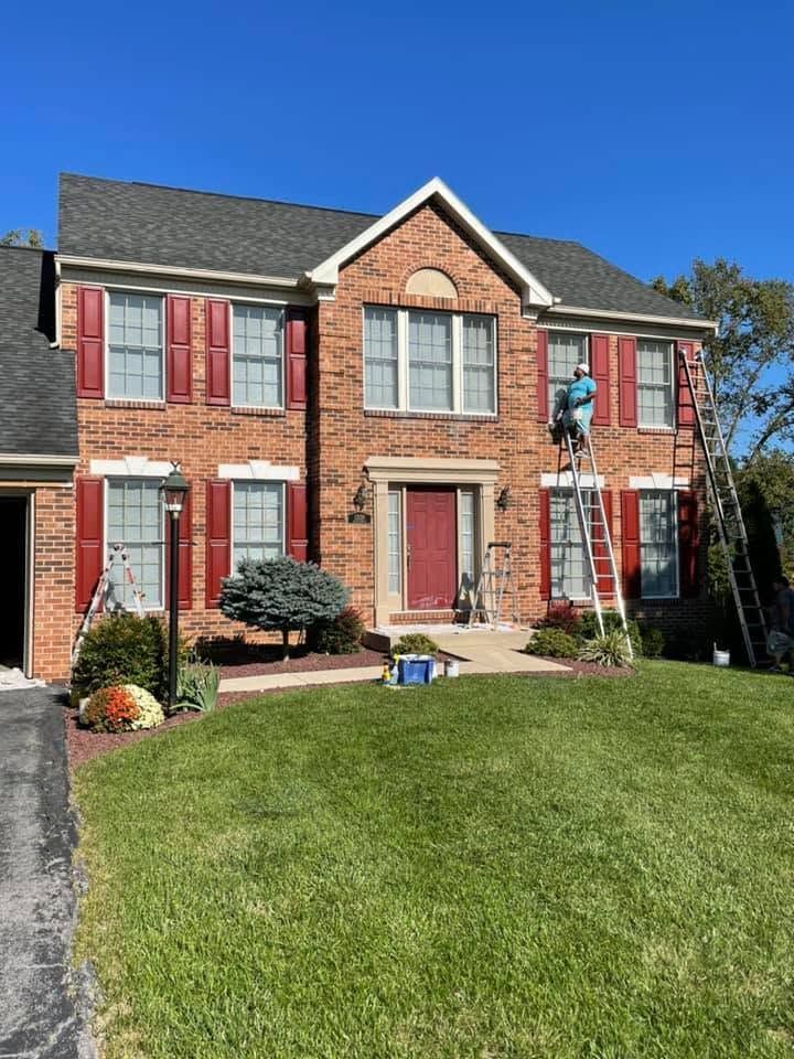 Two-story brick house with red shutters, a front lawn, and a worker on a ladder near the right window.