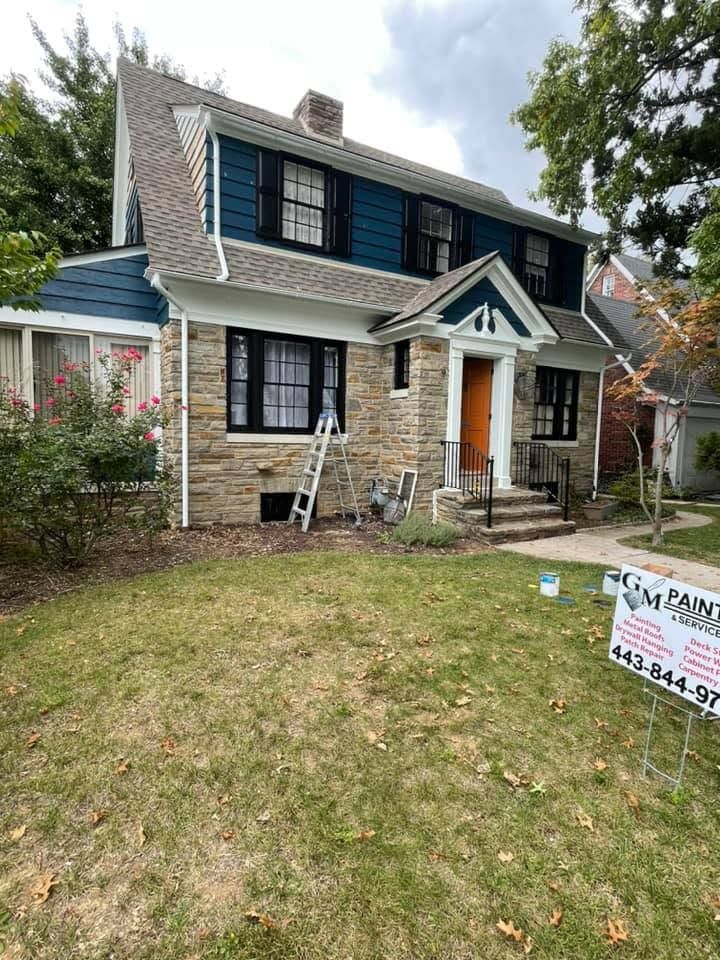 Two-story house with stone facade, dark shutters, and a ladder in the front yard under a cloudy sky