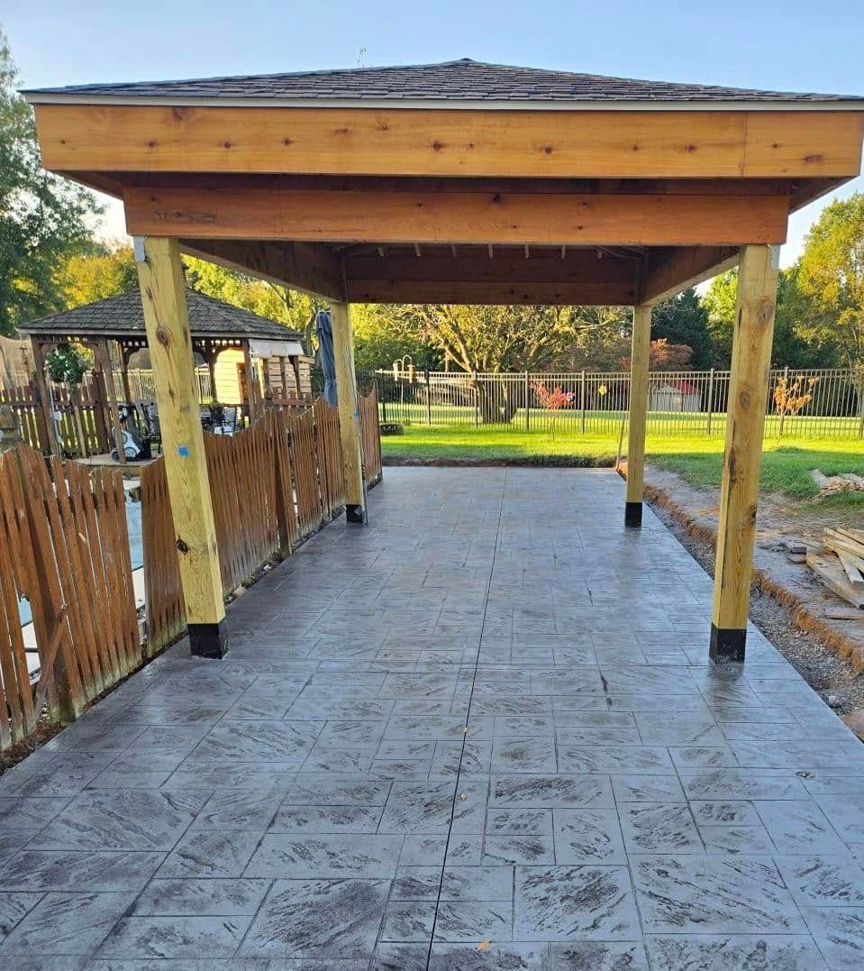 Covered outdoor patio with wooden posts, stone floor, and fenced sides overlooking a grassy yard