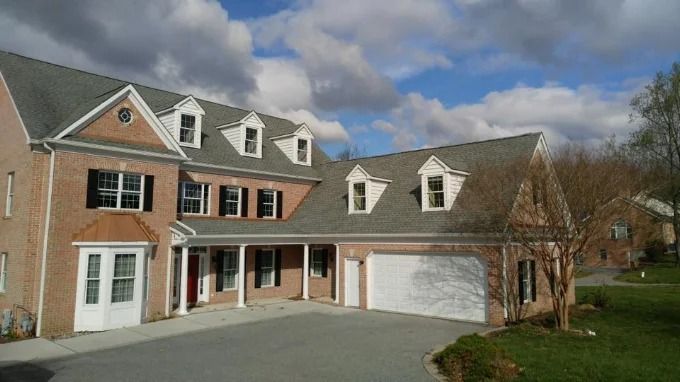 Large brick suburban house with white trim, dormer windows, and attached garage under a cloudy sky