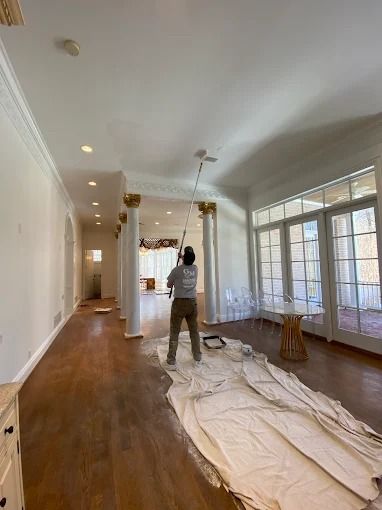 Bright living room with white walls, hardwood floors, large windows, and a person painting near a covered floor.