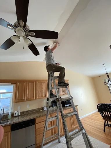Man standing on a ladder adjusting a ceiling light in a kitchen with wood cabinets and a ceiling fan