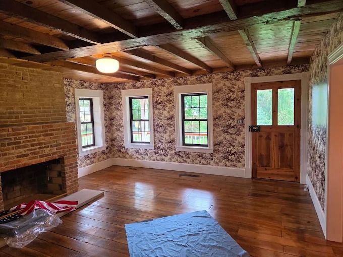 Empty rustic living room with wood floors, exposed beams, brick fireplace, patterned walls, and green-trimmed windows