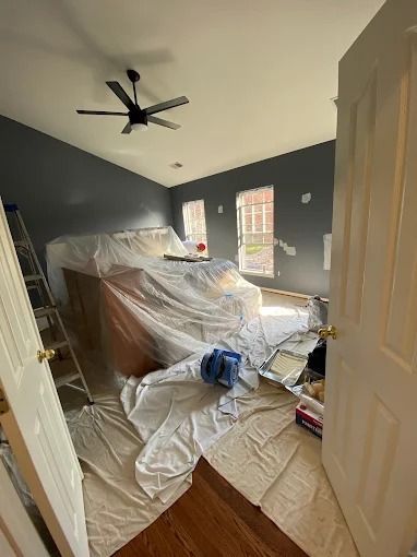 Cluttered bedroom under renovation, with furniture covered in plastic and tools scattered on the floor.