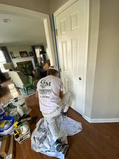 Worker kneeling by a white door, painting trim in a room with a hardwood floor and painting supplies.