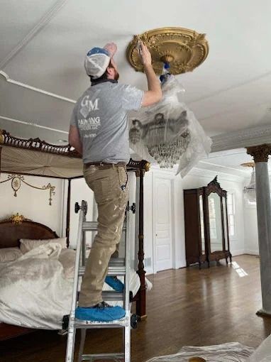 Two workers installing an ornate ceiling medallion in a bedroom with a chandelier and four-poster bed
