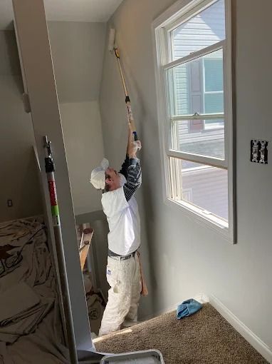 Person painting a wall with a roller beside a window in a room under renovation