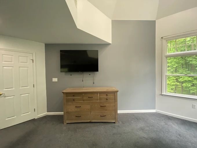 Empty gray bedroom with wall-mounted TV, wooden dresser, white door, and large window.