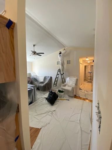Hallway view into a room mid-renovation with plastic sheeting, ladder, covered furniture, and ceiling fan.