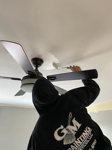 Person reaching up to adjust a black ceiling fan in a room with white ceiling and walls.