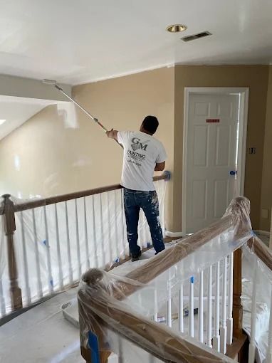 Person painting an upstairs hallway with a roller near a white door and staircase railing.