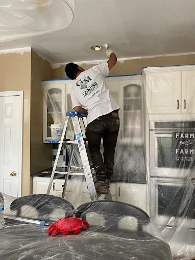 Worker on a ladder painting a ceiling light fixture in a kitchen, with cabinets covered in plastic.