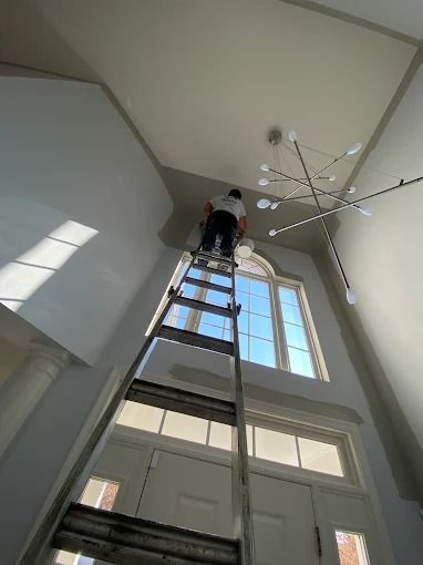 Person on a ladder high in a tall room with large windows and a modern chandelier