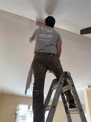Person on a ladder painting a ceiling in a room with a window.
