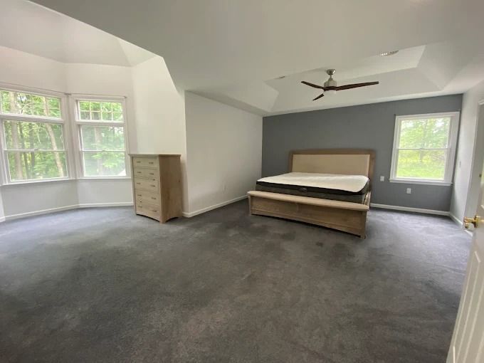 Spacious bedroom with gray carpet, bed, dresser, windows, and ceiling fan in neutral tones