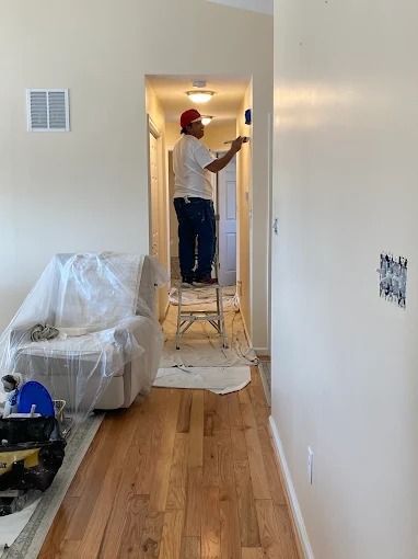 Person painting a hallway wall in a home, with furniture covered in plastic and tools on the floor.