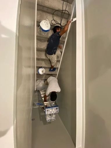 Two workers painting a stairwell, one on a ladder and one on the stairs, with buckets and tools nearby.