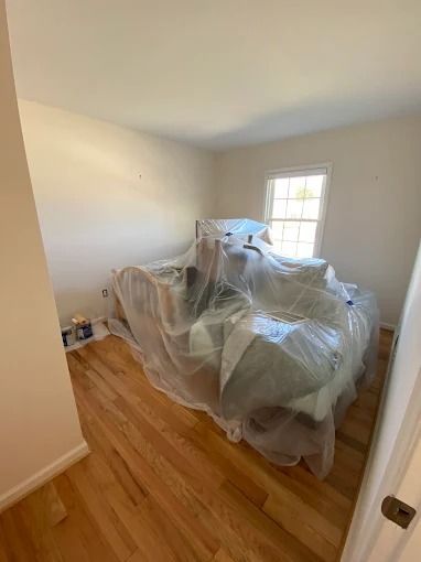 Small room with covered furniture under plastic sheeting, white walls, hardwood floor, and a window.