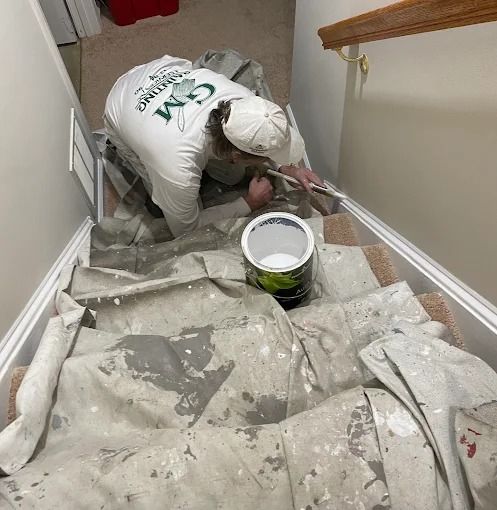 Worker painting stairwell, crouched on carpeted stairs beside a paint bucket.