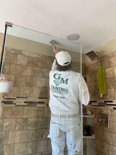 Person in a white cap and hoodie cleaning a glass shower enclosure in a tiled bathroom