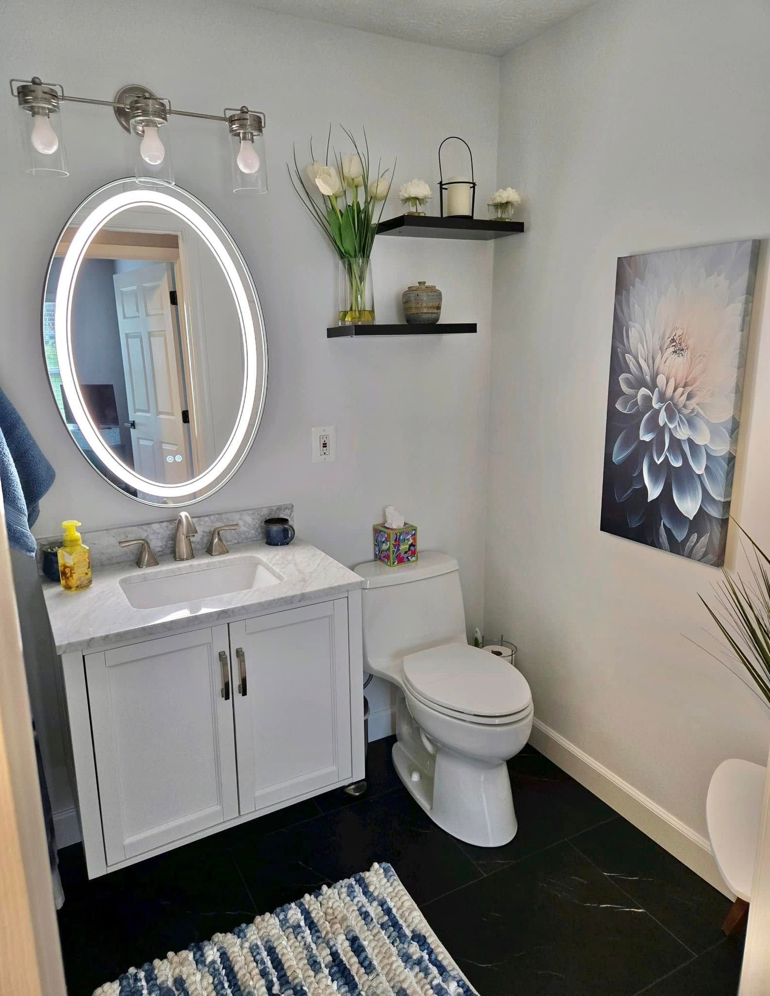 Bright bathroom with white vanity, round lit mirror, toilet, shelves, black floor, and floral wall art.