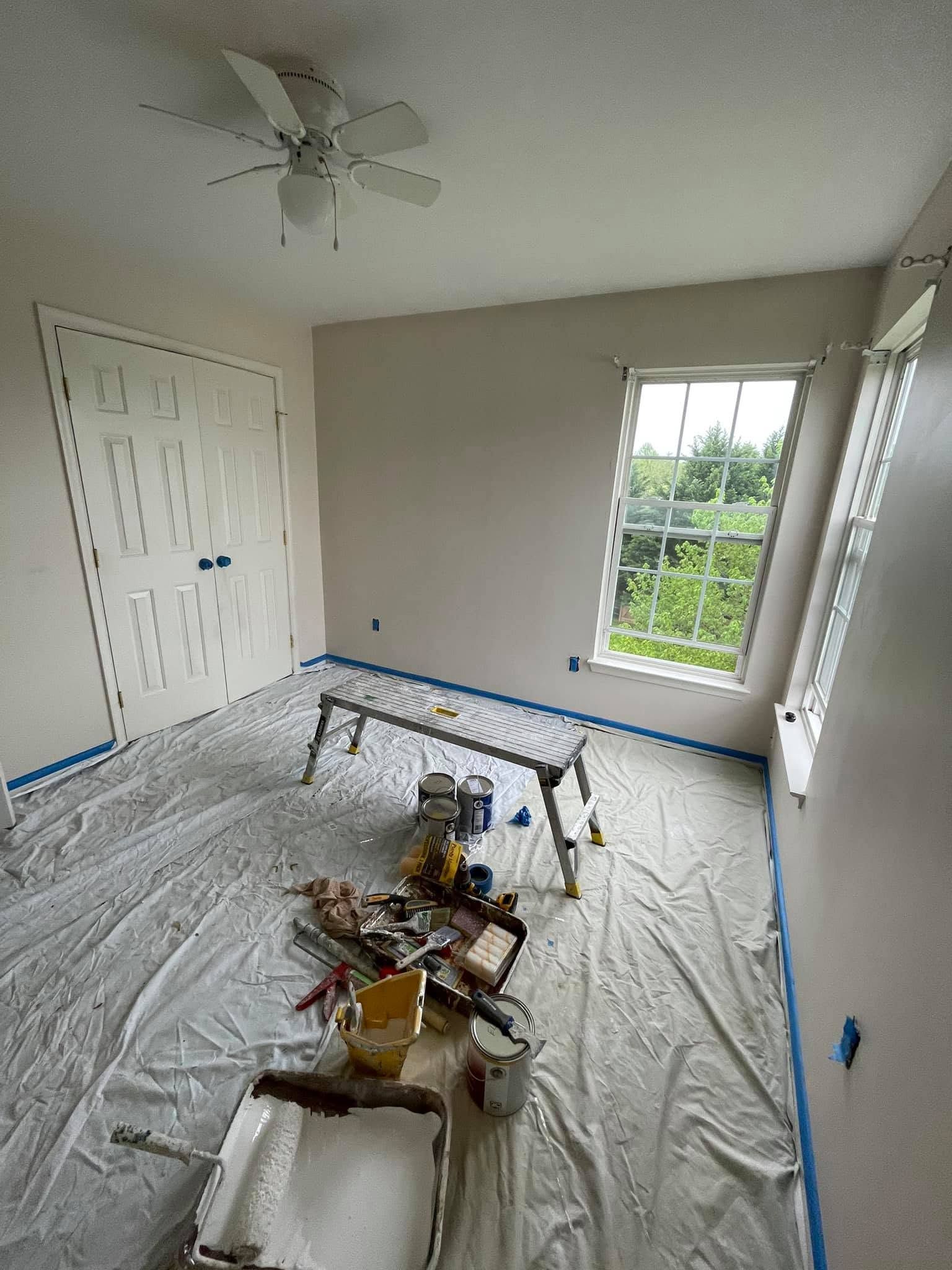 Empty room under renovation with plastic-covered floor, tools, and a ceiling fan near a window.