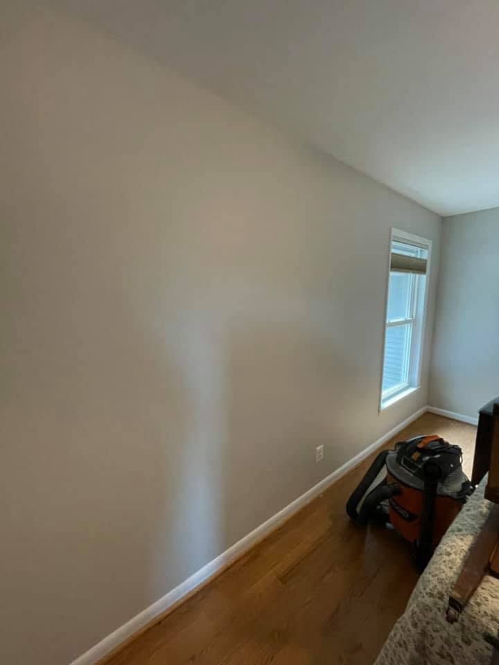 Empty beige hallway with white trim, hardwood floor, and a window; cleaning tools sit on the floor.