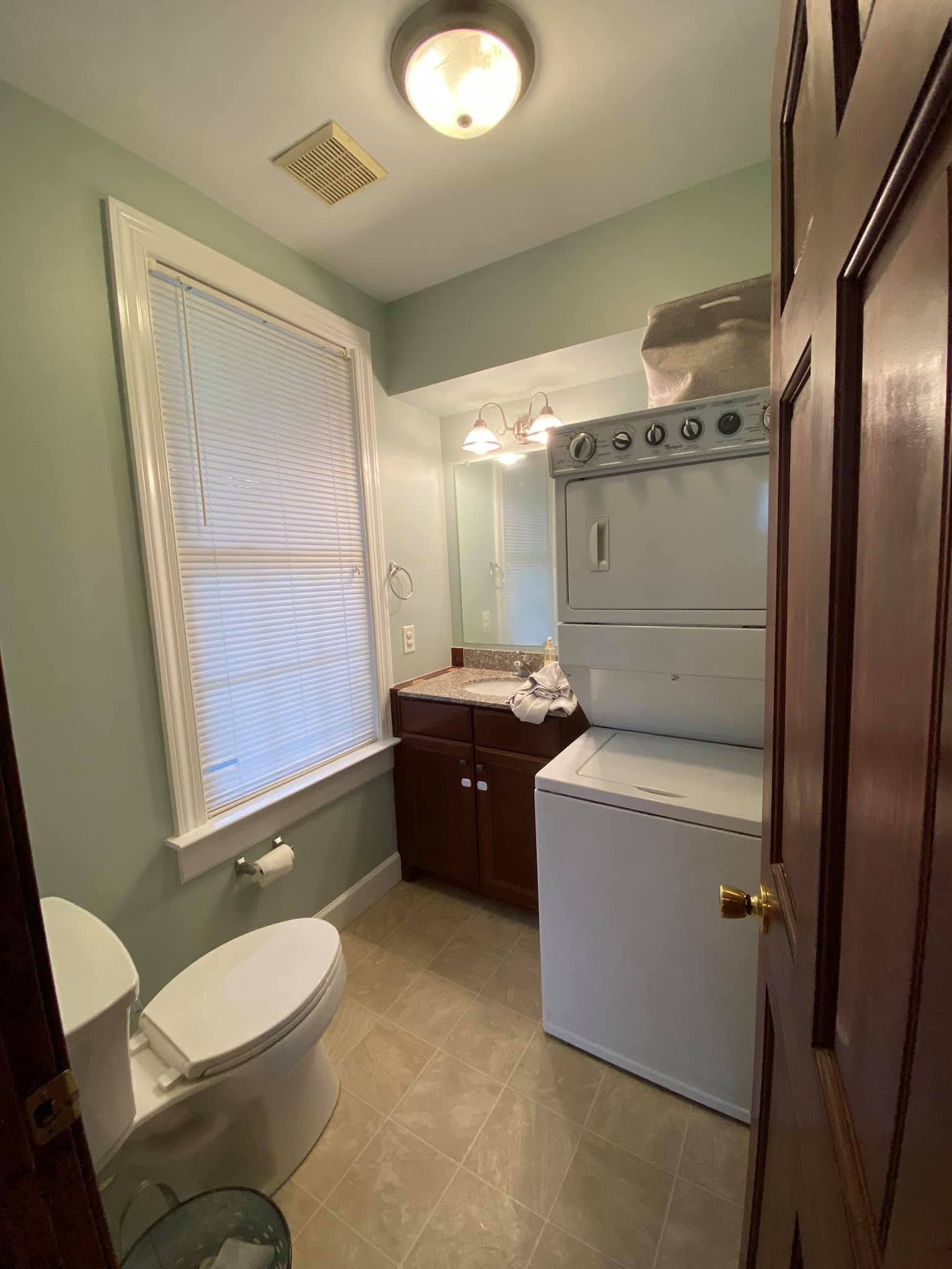 Small bathroom with toilet, sink vanity, stacked washer-dryer, and window with blinds.