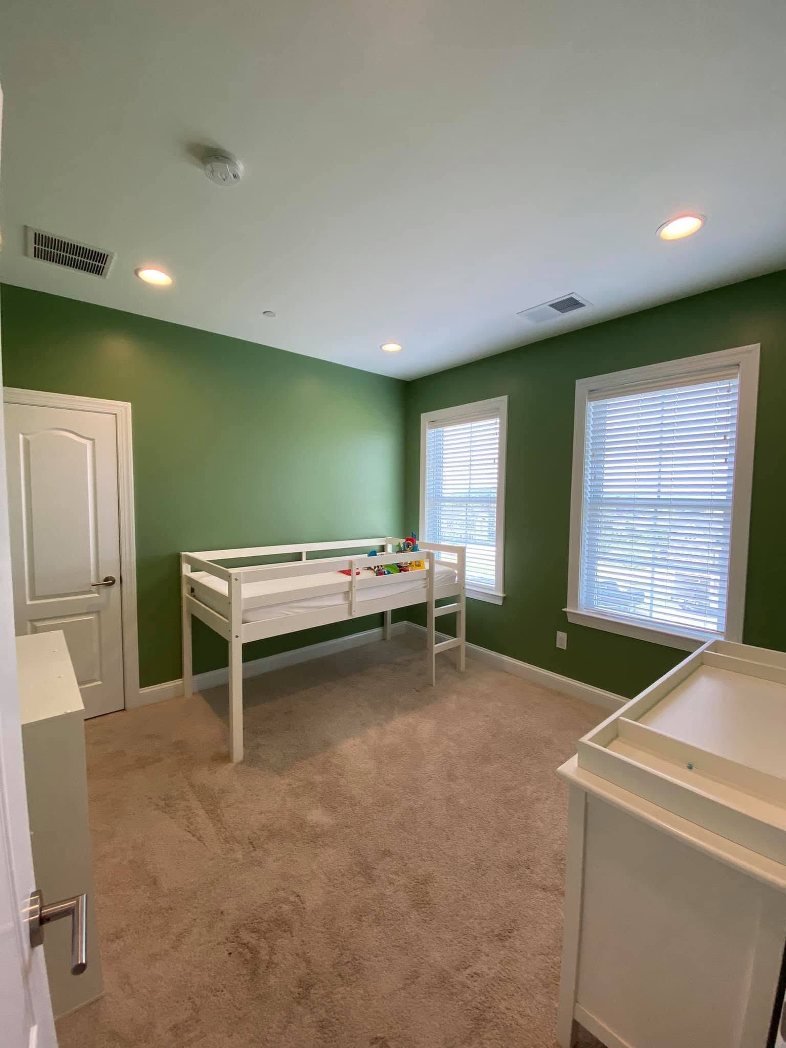 Empty nursery with green walls, white crib, beige carpet, and two bright windows with blinds.