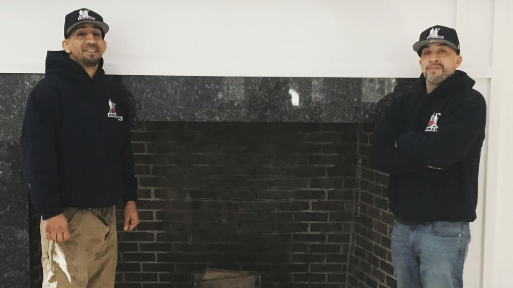 Two men in black jackets and caps standing beside a dark brick fireplace mantle
