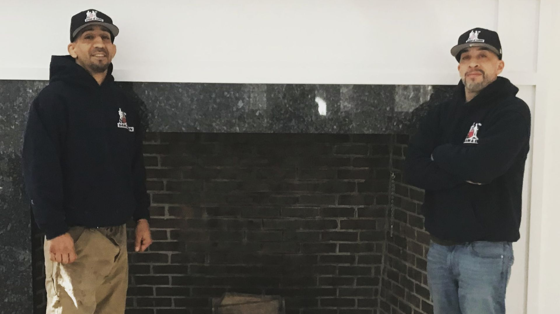 Two men in black jackets and caps standing beside a dark brick fireplace mantle