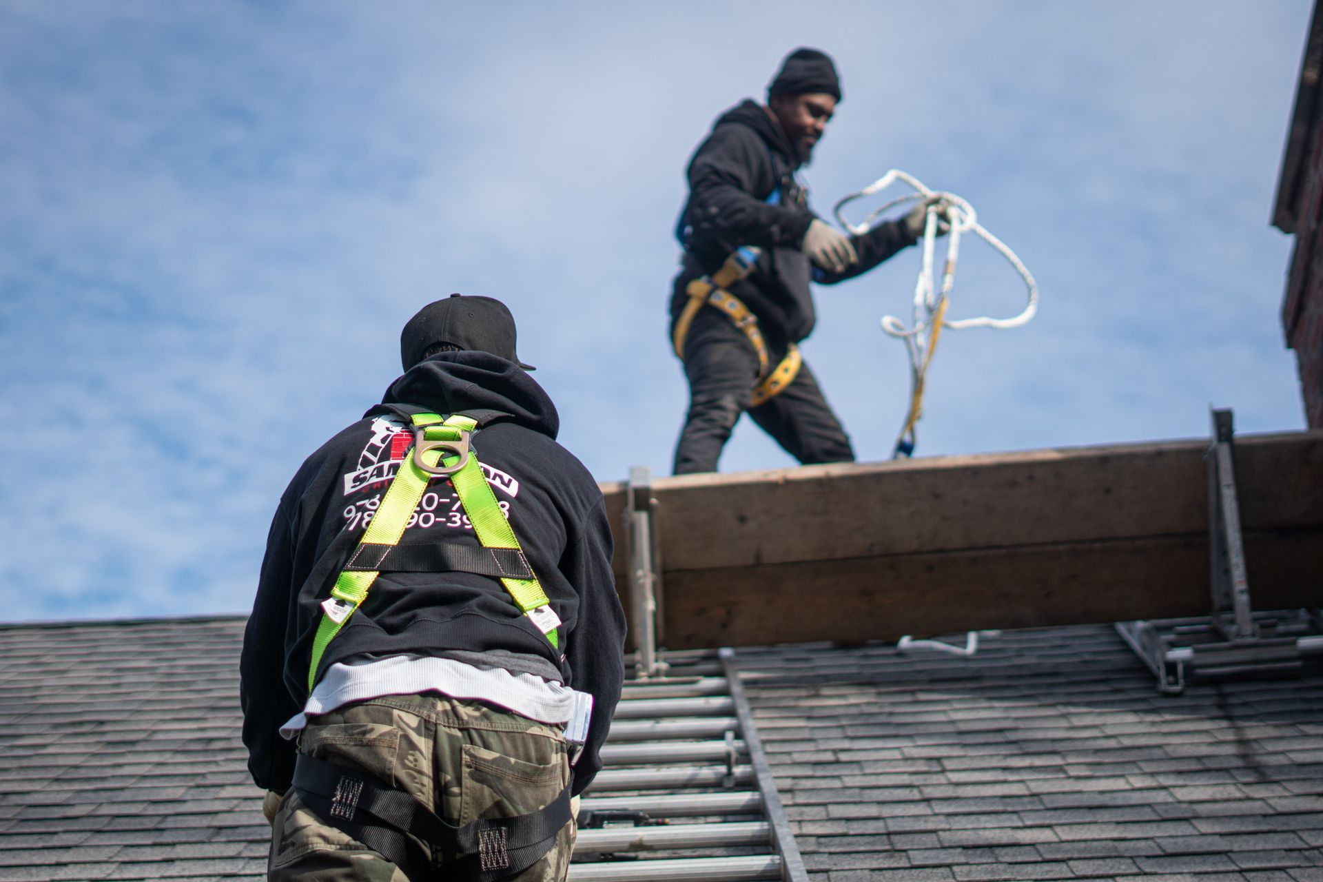 Two workers on a roof using safety harnesses, one standing near the edge and one climbing a ladder.