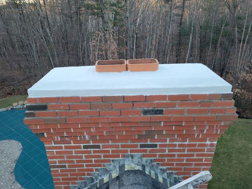 Brick chimney with a white cap and two bricks on top, set against trees outdoors