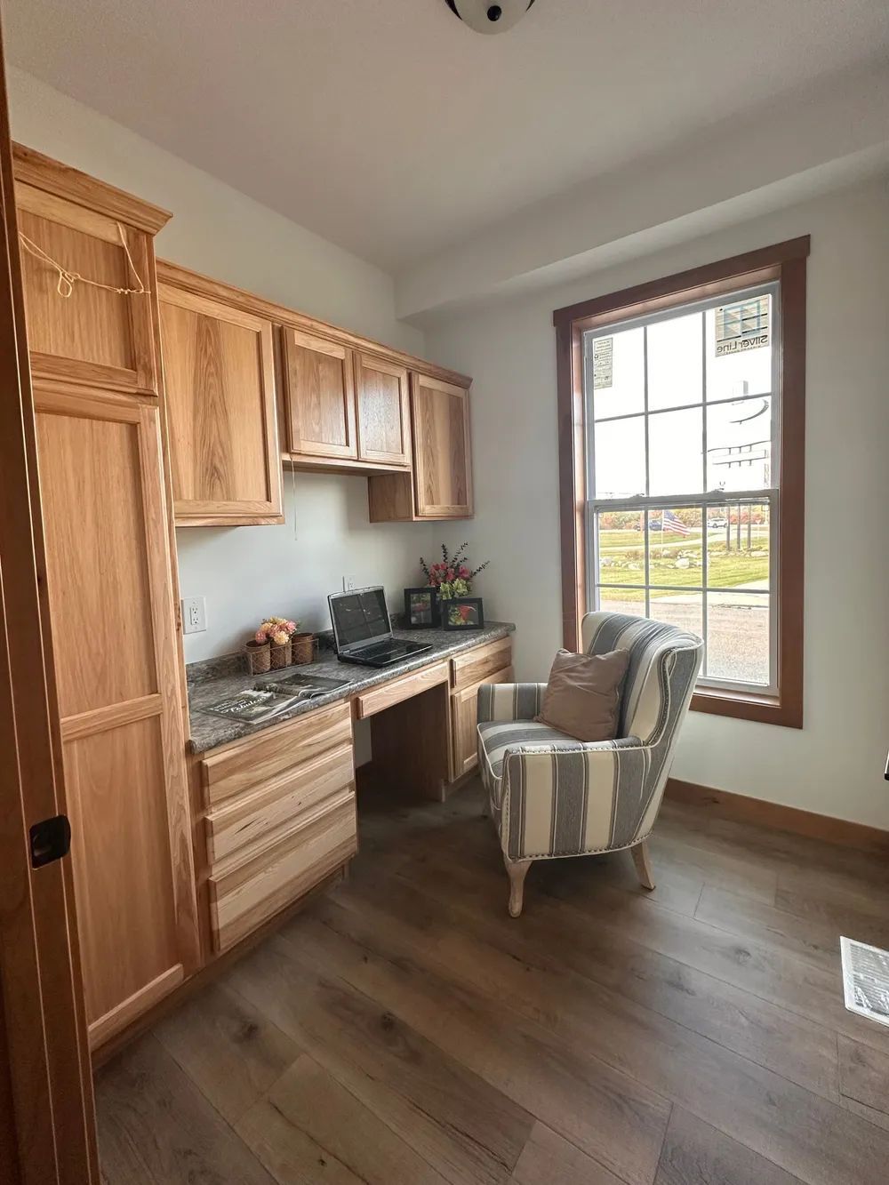 Home office corner with wooden desk, cabinets, striped chair, and a large window.