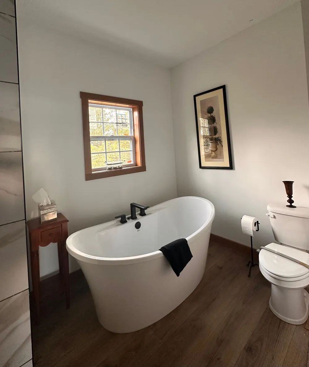 Bright bathroom with a freestanding white tub, toilet, window, and wood floor.