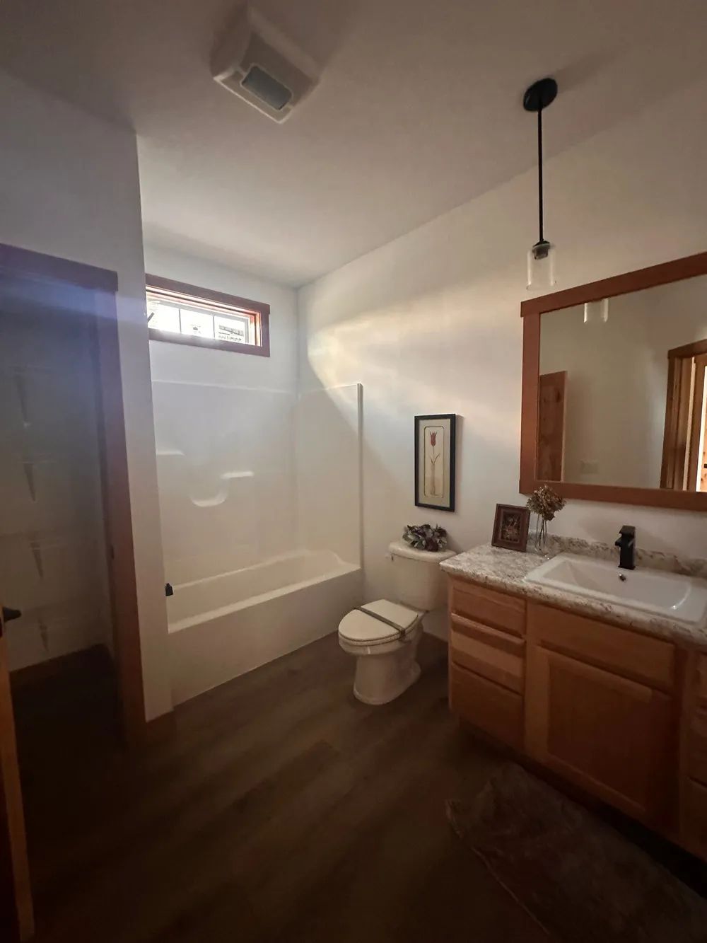 Small bathroom with a white tub, toilet, wooden vanity, and mirror on dark wood flooring.
