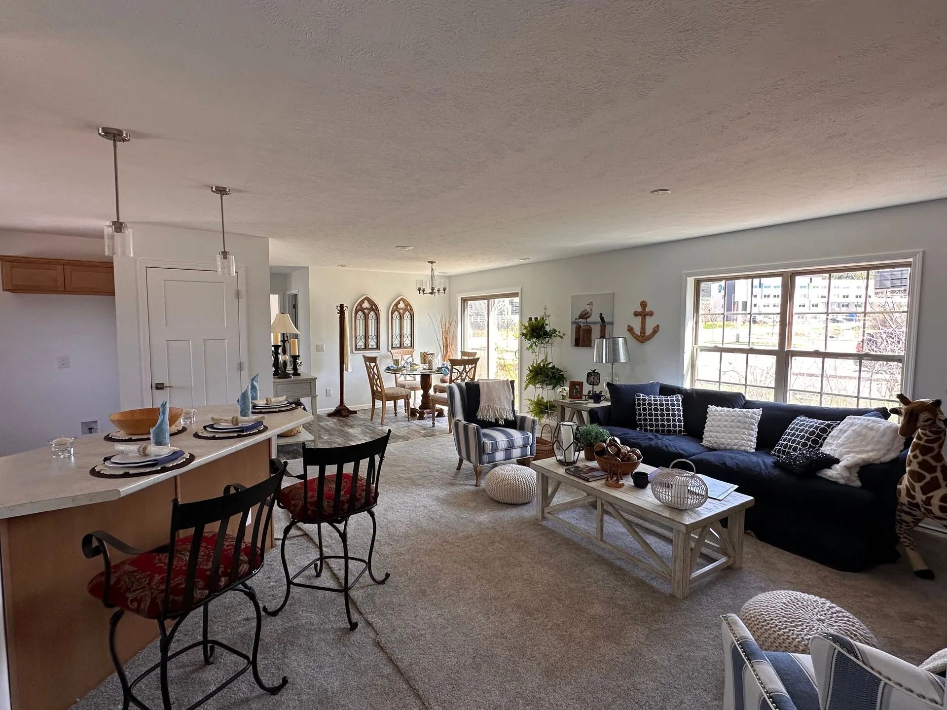 Open-concept living room and kitchen with black sofa, white coffee table, bar stools, and large windows.