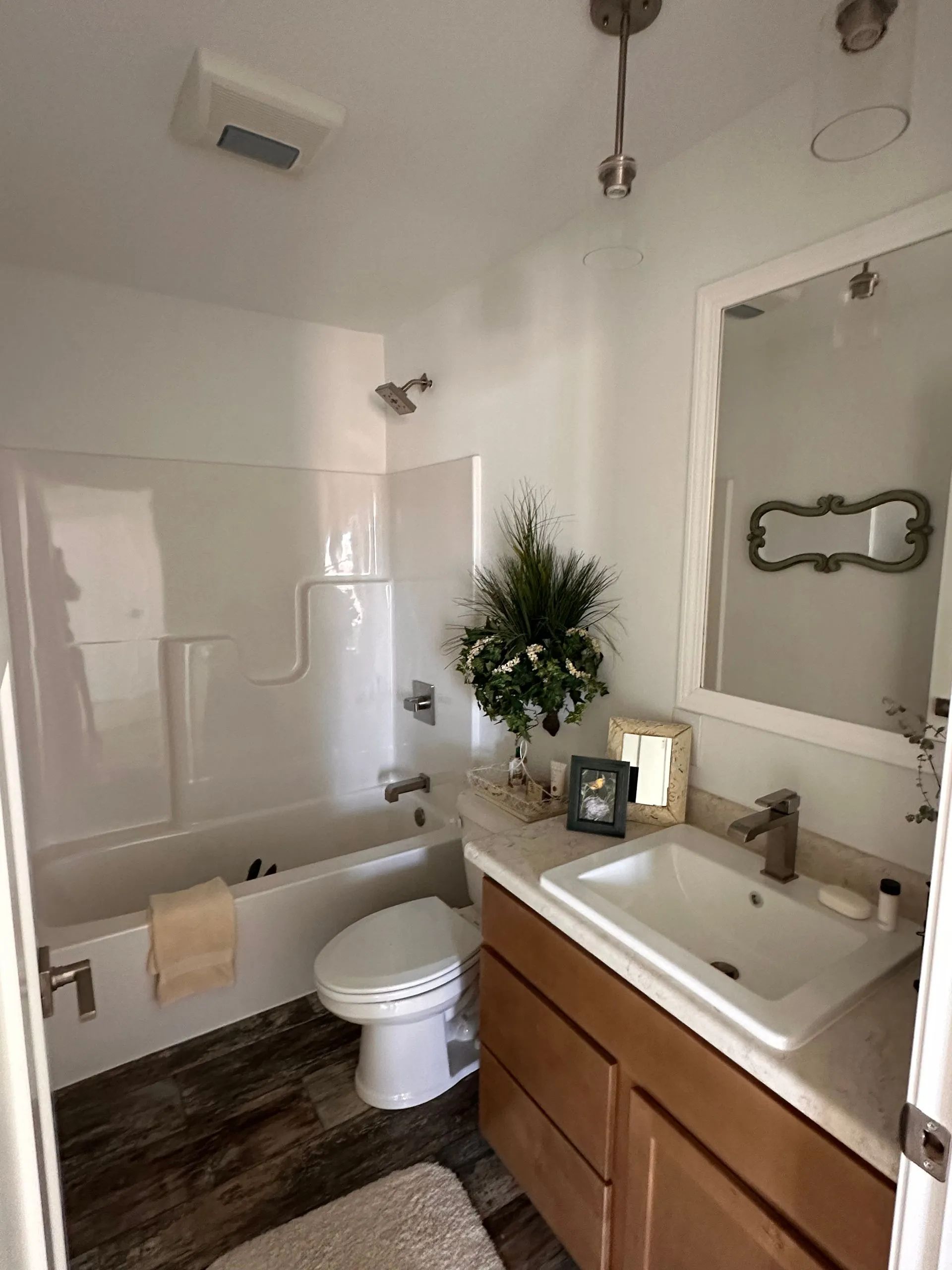 Small bathroom with white tub, toilet, wood vanity, and a potted plant by the sink.