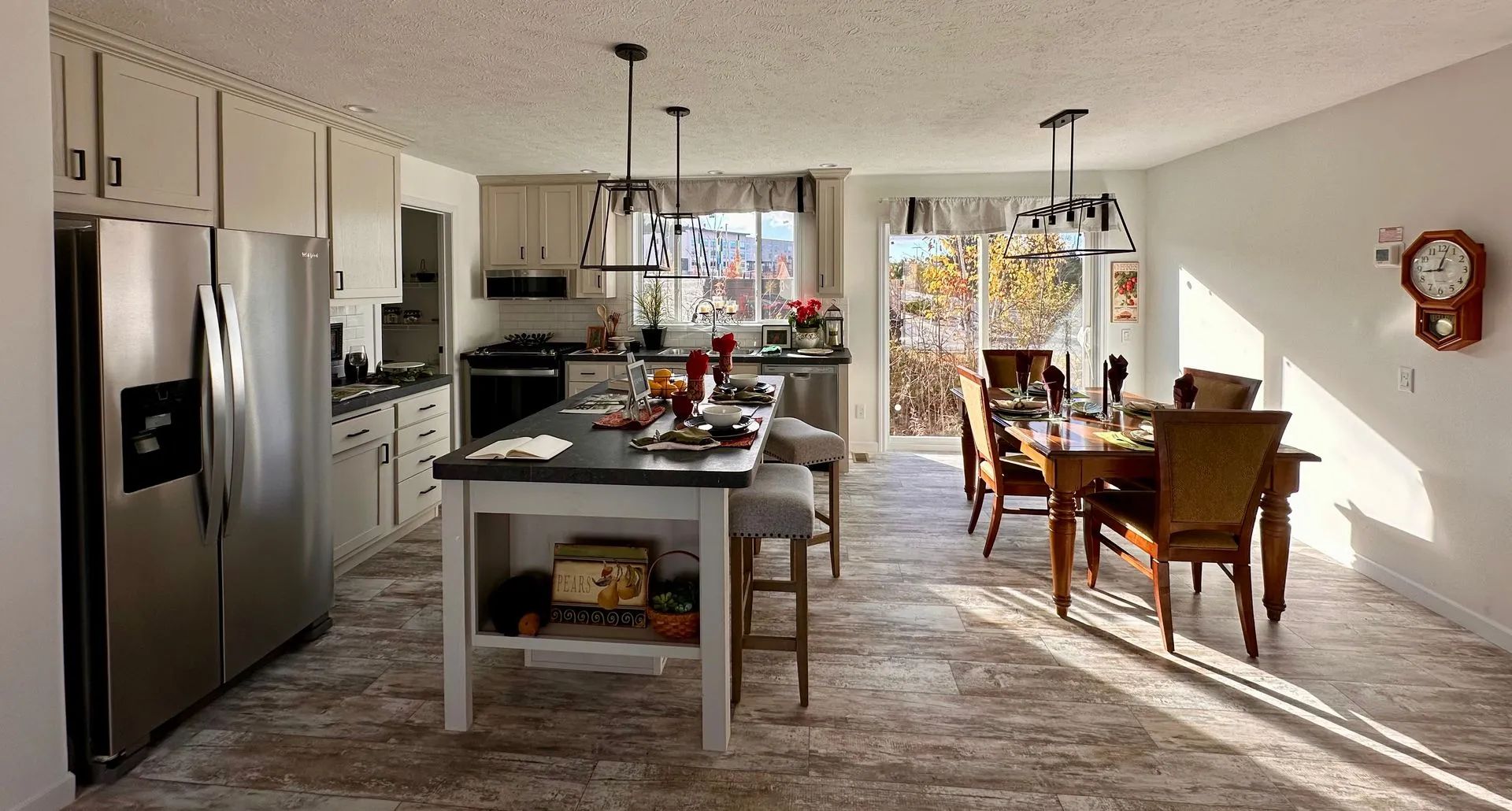 Open-concept kitchen and dining room with island, stainless fridge, wooden table, and sunlight streaming in.