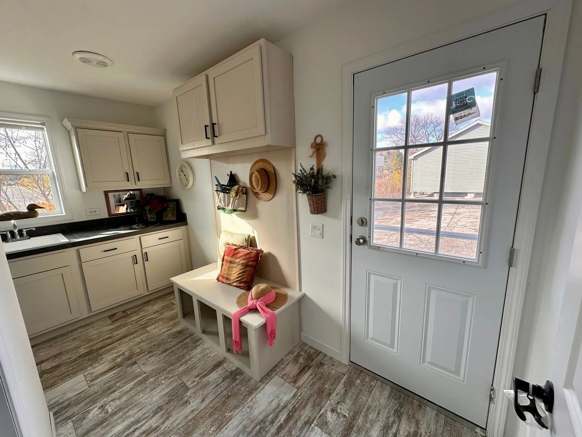 Compact kitchen with white cabinets, wood-look floor, bench with decor, and a glass-paneled door.