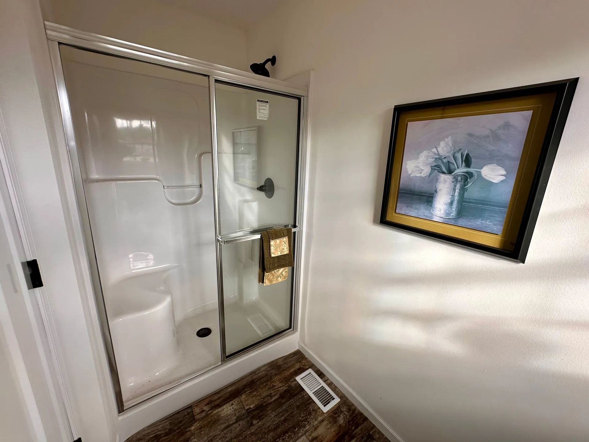 Small shower with glass door and gold handle beside a framed painting on a beige wall.