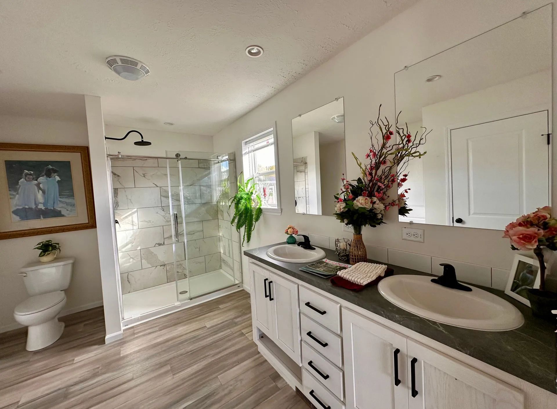 Bright bathroom with double vanity, glass shower, toilet, wood floors, and floral decor