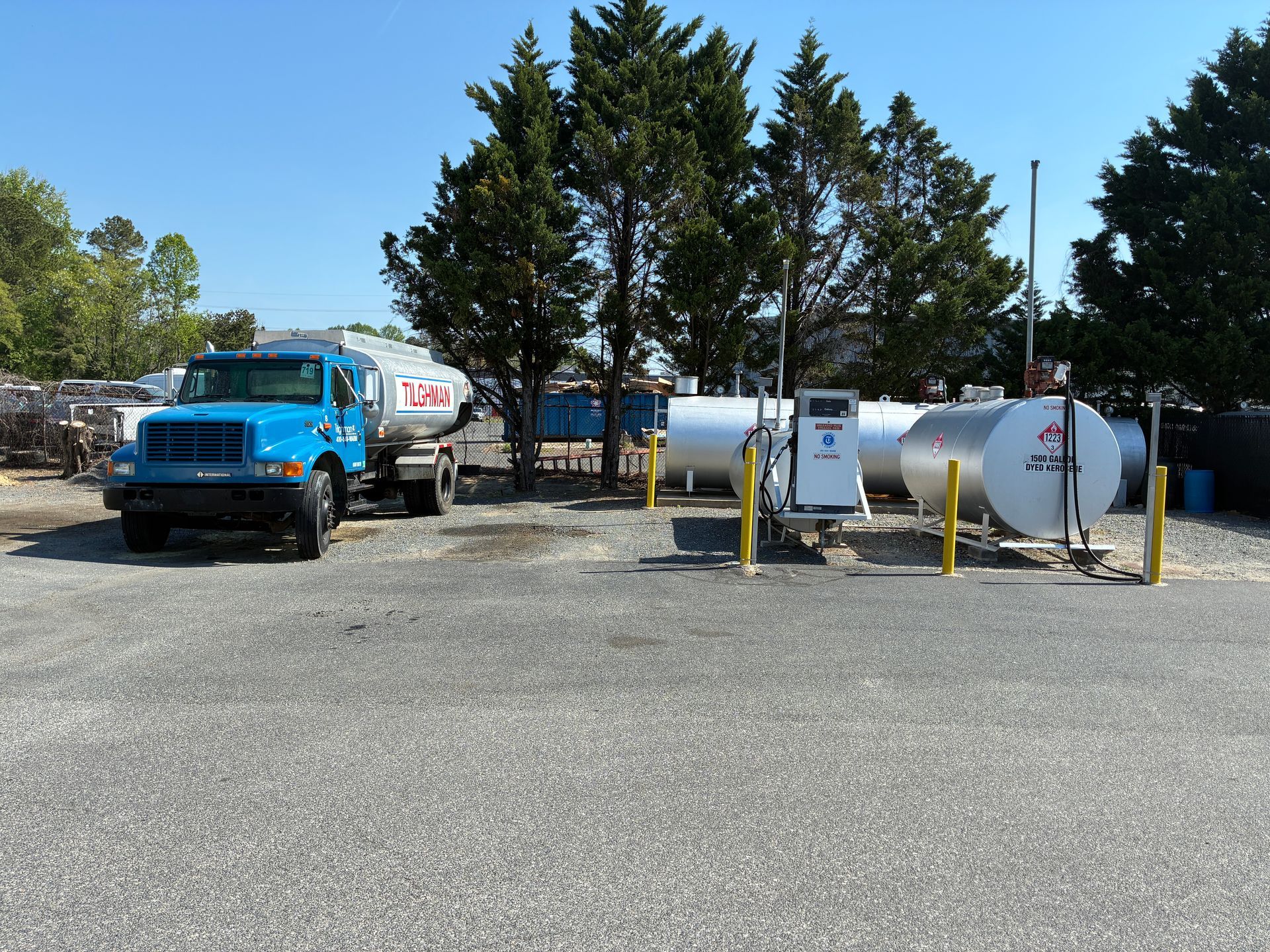 Blue truck beside white tank trailers in a gravel lot with trees and yellow bollards