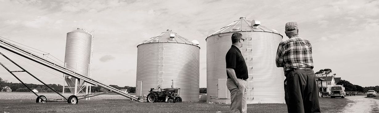 Two people walking beside large grain silos and farm machinery in a rural industrial yard