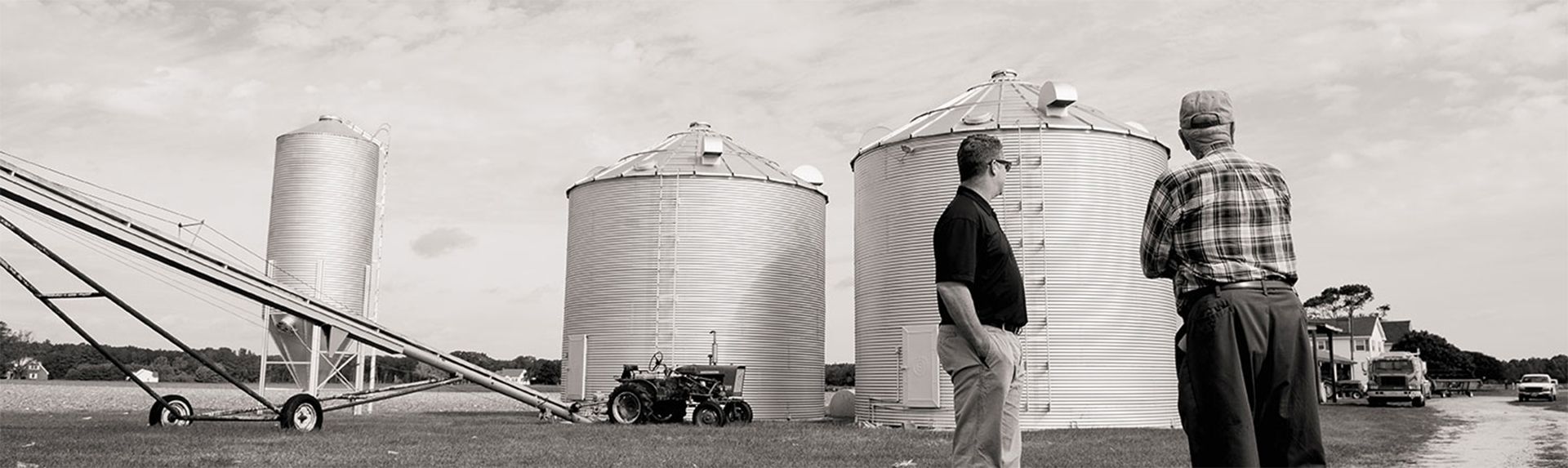 Two people walking beside large grain silos and farm machinery in a rural industrial yard