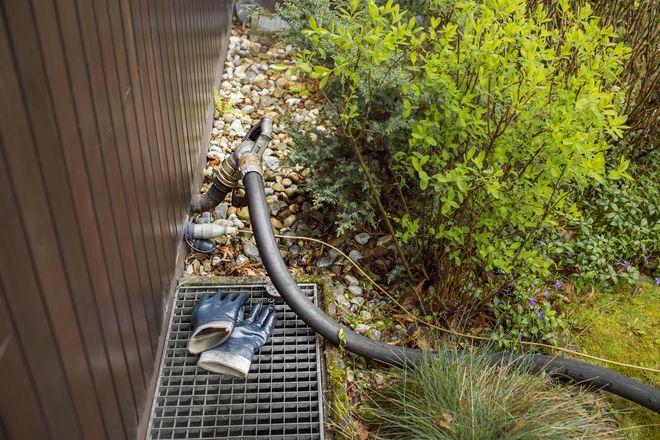 Black cat on a metal grate beside a house, near a hose and shrubs.