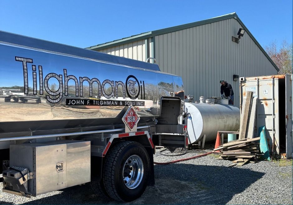Silver Tighman Oil tank truck parked beside a gray building, connected to a hose for fuel delivery.
