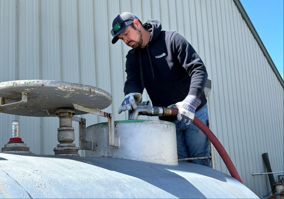 Worker in a hard hat uses a hose to inspect or clean a large metal tank outdoors.