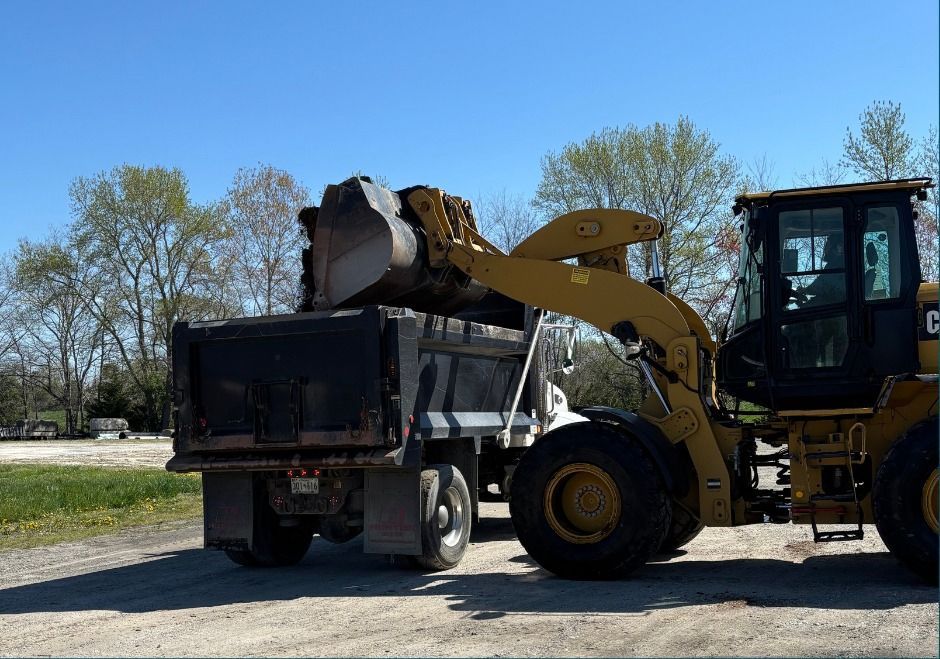 Yellow front loader dumping dirt into a black dump truck on a sunny gravel lot.