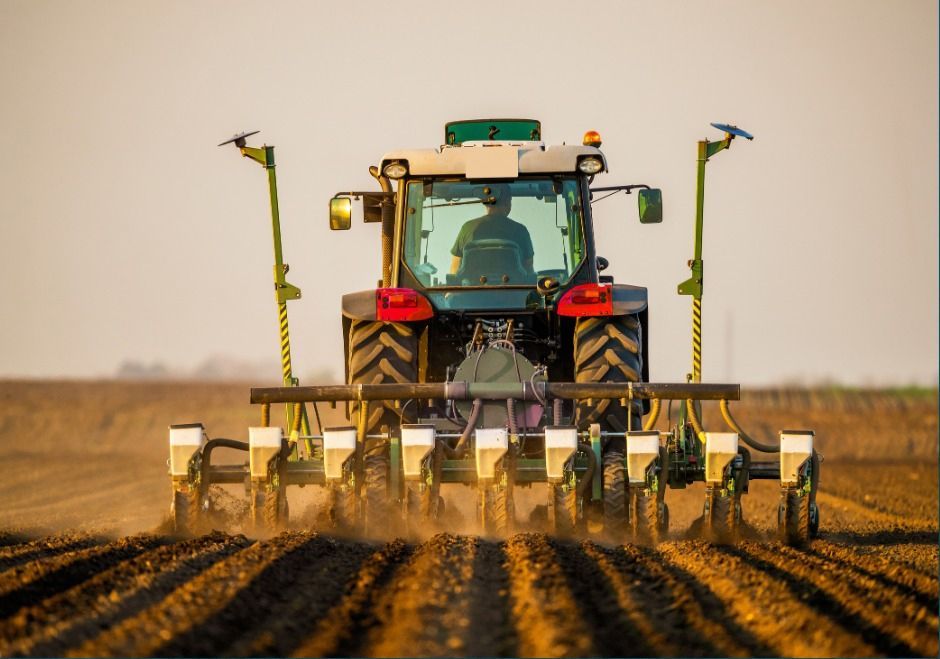 Tractor planting crops in a dusty field, seen from behind with rows of furrows and sprayers extended.